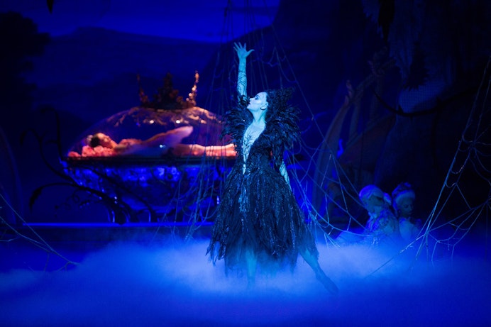 Alice Topp as Carabosse, arm raised, by a sleeping figure under blue light.
